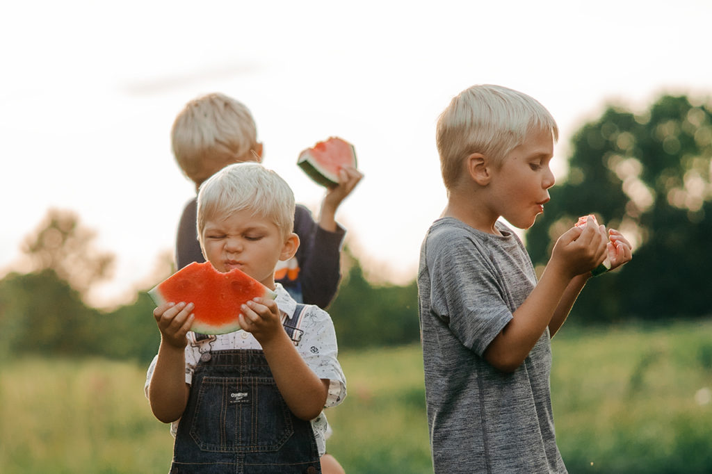 Three boys eating watermelon in a filed while one makes a silly sour face.